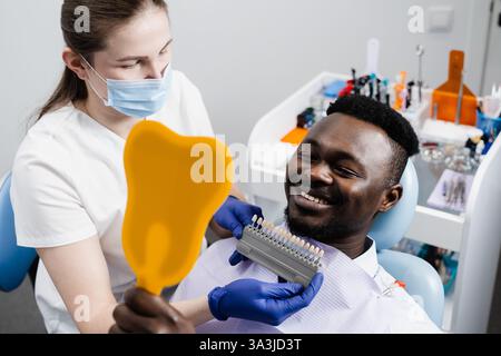African est à la recherche de facettes ou d'implants de dents de couleur assortie d'échantillons dans les mains du médecin. Odontologie. Dentiste montrant les dents guide de nuances de couleur pour la dent Banque D'Images