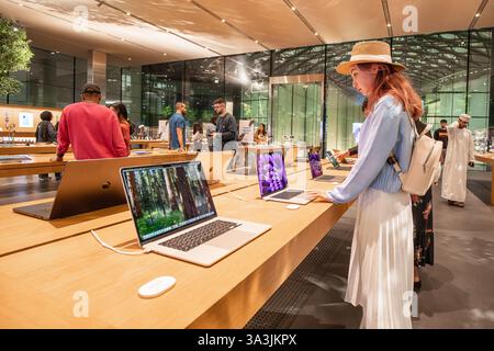 17 janvier 2025, Abu Dhabi, Émirats arabes Unis : jeune femme essayant un ordinateur portable dans un magasin d'électronique Apple moderne, avec d'autres clients naviguant sur des produits Banque D'Images