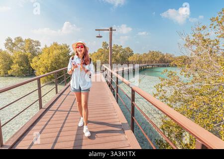Femme excitée dans un chapeau de soleil tenant un smartphone sur une passerelle en bois à travers des mangroves vertes luxuriantes Banque D'Images