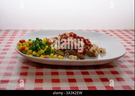 Une assiette de pâtes cuites mélangées à de la viande hachée est accompagnée d’un côté coloré de pois verts, de maïs et de poivrons rouges, le tout servi sur un damier classique Banque D'Images
