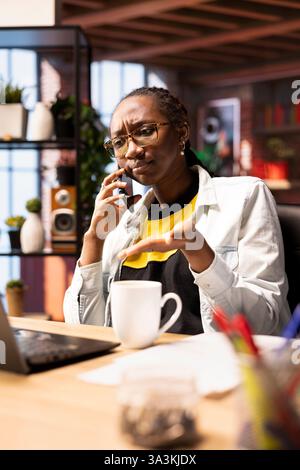 Télétravailleur au bureau à domicile bouleversé par les mauvaises nouvelles reçues lors d'un appel téléphonique. Femme afro-américaine agacée recevant un appel téléphonique frustrant sur smartphone d'un collègue de travail Banque D'Images