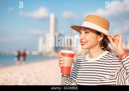 Jeune femme portant un chapeau de paille tenant une tasse de café à emporter assise sur une plage avec l'horizon d'Abu Dhabi en arrière-plan Banque D'Images