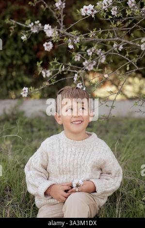 Un garçon est assis sous un amandier en fleurs, cueillant des amandes fraîches et les tenant dans ses mains. Banque D'Images