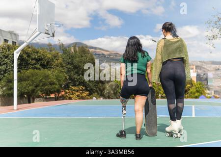 Deux femmes, dont une avec une jambe prothétique, se tiennent debout sur un terrain de basket-ball avec une planche à roulettes et des patins à roulettes, profitant de la vue Banque D'Images