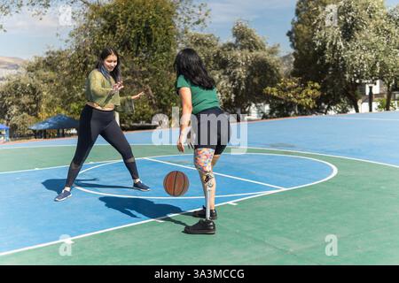 Deux jeunes femmes jouant au basket-ball par une journée ensoleillée, dont une avec une prothèse de jambe montrant détermination et inclusivité dans le sport Banque D'Images