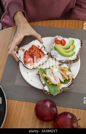 Une femme gracieuse, élégamment habillée, profite d'un petit déjeuner raffiné composé d'un trio de toasts au fromage à la crème : l'un garni de tranches de tomates fraîches, ano Banque D'Images
