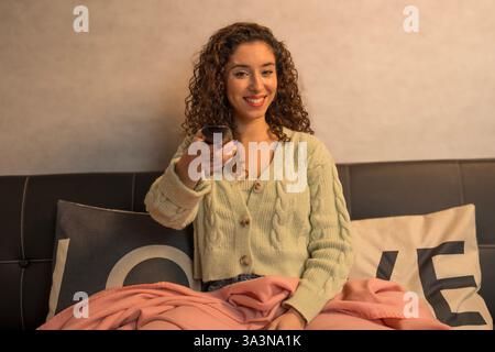Jeune femme assise confortablement sur un canapé sous une couverture, changeant de chaînes avec télécommande et profitant de son émission de télévision préférée Banque D'Images