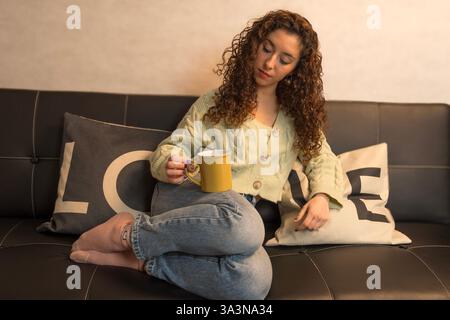 Jeune femme sereine profitant d'un moment de calme sur un canapé confortable, tenant une tasse et entourée d'oreillers sur le thème de l'amour Banque D'Images