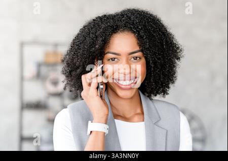 Une femme d’affaires afro-américaine au sourire radieux parle sur son smartphone dans un bureau lumineux et moderne. Une femme professionnelle engagée dans une conversation d'affaires agréable Banque D'Images