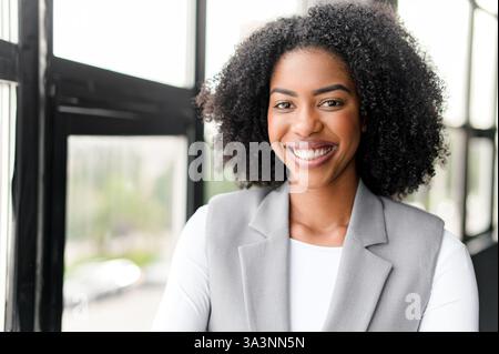 Une femme d'affaires afro-américaine rayonnante avec un sourire éclatant se tient confiante dans un bureau aux grandes fenêtres, symbolisant l'ouverture et un environnement de travail positif. Banque D'Images