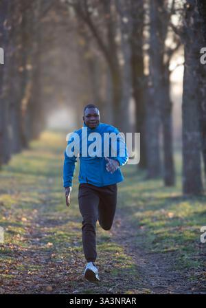 Homme en veste bleue jogging le long du chemin bordé d'arbres au petit matin, profitant de l'air frais et de la lumière au lever du soleil. Sport et mode de vie sain conc Banque D'Images