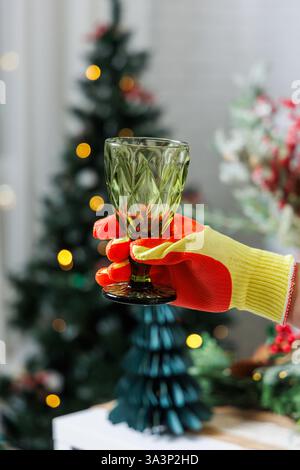 Une personne vêtue de gants rouges et jaunes vifs tient gracieusement un verre à vin élégant devant un sapin de Noël magnifiquement orné Banque D'Images