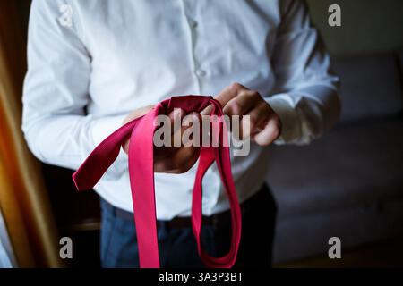 Un homme vêtu d'une chemise blanche impeccable attache habilement une cravate rouge vif autour de son cou, se concentrant sur la formation parfaite du noeud Banque D'Images