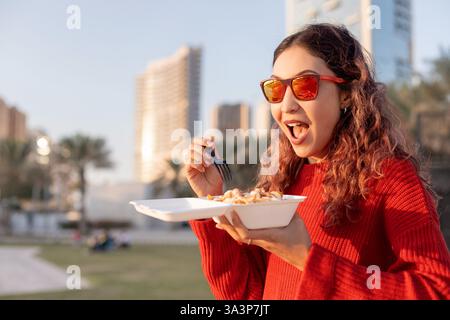 Heureuse jeune femme appréciant la nourriture de rue dans un parc de la ville, tenant une boîte à emporter et savourant un délicieux repas Banque D'Images