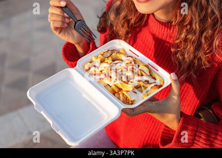 Gros plan d'une femme dégustant des frites chargées garnies de poulet, ketchup et mayonnaise, servies dans un récipient blanc à emporter Banque D'Images