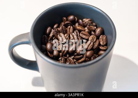 Gros plan d'une tasse en céramique noire remplie de grains de café torréfiés, placée sur une surface blanche. Banque D'Images