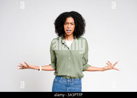 Une femme afro-américaine se tient debout avec une expression incrédule, ses bras écartés dans un geste de questionnement, sur un fond blanc propre. L'image dépeint un moment de confusion ou d'incrédulité Banque D'Images