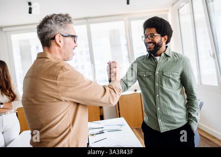 Deux professionnels se serrant la main dans un bureau moderne lumineux, scellant un accord commercial. Les membres de l'équipe souriants en arrière-plan renforcent la confiance, l'accord et une atmosphère de travail positive. Banque D'Images