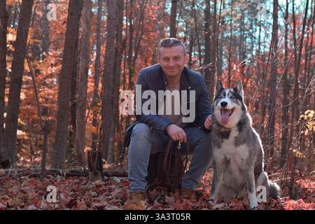Homme d'âge moyen assis sur une souche d'arbre dans une forêt d'automne avec un chien Husky et regardant la caméra souriant Banque D'Images