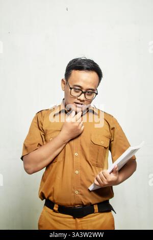 sage professeur asiatique indonésien debout tenant le livre isolé sur fond blanc avec chemise kaki Banque D'Images