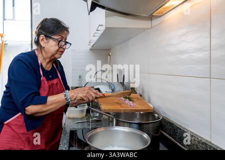Femme latine âgée portant un tablier préparant des ingrédients sur une planche à découper tout en cuisinant dans sa cuisine à la maison Banque D'Images