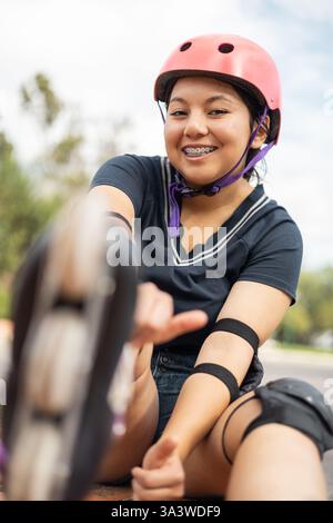 Une jeune fille joyeuse s'assoit, enfilant ses patins avant de commencer à patiner, portant un casque, des genouillères et des coudières pour plus de sécurité. Banque D'Images