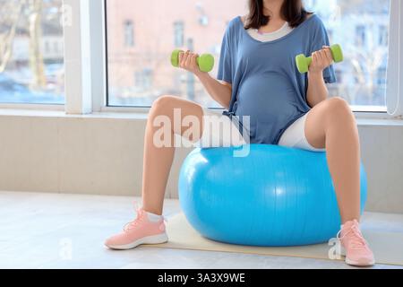 Jeune femme enceinte s'entraînant avec fitball et haltères près de la fenêtre à la maison Banque D'Images