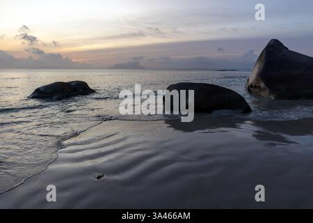 Une scène de plage paisible avec du sable lisse, de grands rochers éparpillés, et un ciel doré coucher de soleil se reflétant sur la surface de l'eau, exsudant calme et con Banque D'Images