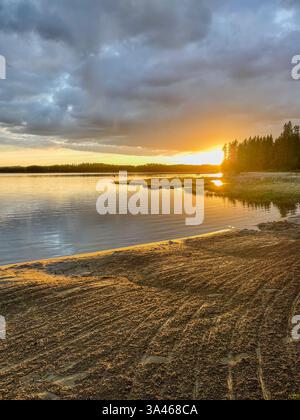 Une vue sur le coucher du soleil depuis un bord de lac sablonneux, avec la lumière dorée illuminant le paysage. La scène comprend une forêt lointaine et des montagnes le long du hor Banque D'Images