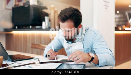Homme, écriture et propriétaire de petite entreprise avec livre sur ordinateur portable pour la gestion de café, rapport ou calendrier. Homme d'affaires, employé ou barista avec des documents ou Banque D'Images