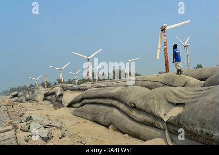 Un homme du coin marche près des éoliennes sur les rives de l’île de Kutubdia. Autrefois symbole d’espoir pour l’énergie verte, ces structures rouillées sont aujourd’hui abandonnées. Une fois opérationnels et produisant de l’électricité, ils sont tombés en délabrement, suscitant des inquiétudes quant à la mauvaise gestion et à l’avenir des initiatives du Bangladesh en matière d’énergies renouvelables. Île de Kutubdia, Bangladesh. Banque D'Images