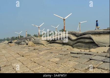 Un homme du coin marche près des éoliennes sur les rives de l’île de Kutubdia. Autrefois symbole d’espoir pour l’énergie verte, ces structures rouillées sont aujourd’hui abandonnées. Une fois opérationnels et produisant de l’électricité, ils sont tombés en délabrement, suscitant des inquiétudes quant à la mauvaise gestion et à l’avenir des initiatives du Bangladesh en matière d’énergies renouvelables. Île de Kutubdia, Bangladesh. Banque D'Images