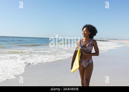 Femme en maillot de bain tenant une planche de surf marchant le long de la plage, profitant de la journée ensoleillée, espace de copie Banque D'Images