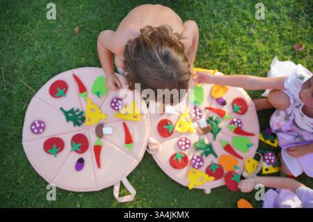 Les enfants se livrent à une activité de plein air ludique en utilisant de fausses pièces colorées de pizza sur l'herbe. La vue de dessus capture la créativité et l'interaction en tant qu'enfants Banque D'Images