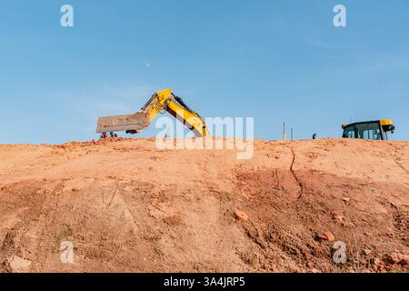 Excavatrice jaune creusant activement de la terre sur un chantier de construction contre un ciel bleu clair. Le bras de la machine s'étend au-dessus du bord de la terre Banque D'Images