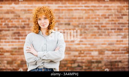 Portrait d'une jeune femme rousse confiante aux bras croisés posant devant un mur de briques Banque D'Images