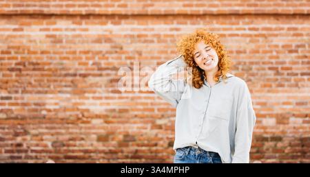 Portrait d'une jeune femme joyeuse aux cheveux roux bouclés, portant une chemise gris clair et un Jean, touchant ses cheveux et souriant les yeux fermés, debout Banque D'Images