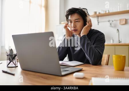 Un jeune homme asiatique étudie attentivement à la maison, absorbé par son cours en ligne et portant des écouteurs. Banque D'Images