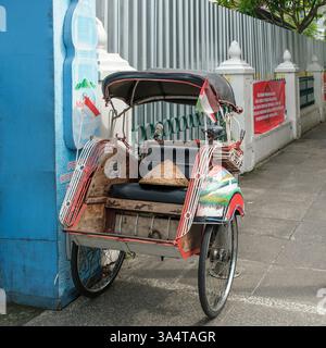 Pousse-pousse traditionnel indonésien, connu sous le nom de becak, garé sur un trottoir à yogyakarta, arborant le drapeau indonésien et un chapeau conique Banque D'Images