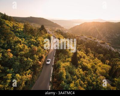 Monténégro, Kotor - 13 juillet 2024 : White Cabriolet roule le long d'une route de haute montagne parmi les sommets boisés. Drone Banque D'Images