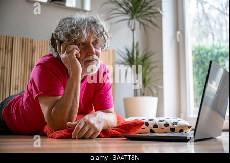 Un homme âgé avec des cheveux gris bouclés et une barbe se trouve sur le sol, parlant au téléphone tout en regardant un ordinateur portable. Il porte une chemise rose vif, appea Banque D'Images
