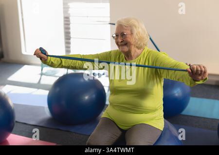 Femme senior exerçant avec la bande de résistance sur la balle de yoga, souriant en classe de fitness Banque D'Images