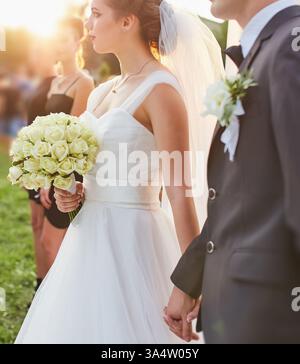 Gros plan d'une mariée tenant un bouquet de rose blanche et la main du marié pendant leur cérémonie de mariage, mettant en évidence l'élégante robe de mariée et le marié Banque D'Images
