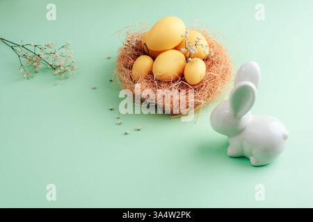 Nid de Pâques avec des œufs jaunes, des fleurs séchées et un lapin blanc en céramique sur un fond vert pastel, symbolisant la célébration du printemps. Concept de Pâques. T Banque D'Images