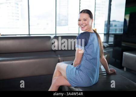 Femme souriante dans un bureau moderne portant une robe décontractée, assise sur un canapé, espace de copie Banque D'Images