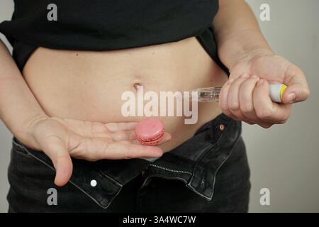 Une femme injecte Ozempic dans son abdomen avec un stylo injecteur tout en tenant un macaron, symbolisant l'alimentation et la perte de poids. Banque D'Images