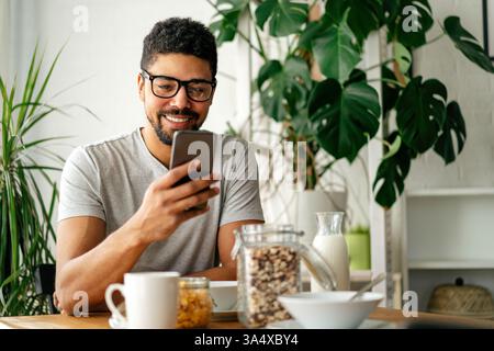 Jeune homme noir mangeant le petit déjeuner et lisant les nouvelles en ligne. Bel homme utilisant un téléphone portable Banque D'Images
