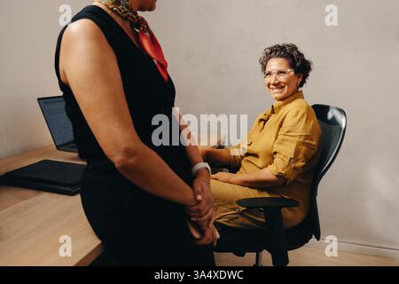 Deux femmes d'affaires ayant une discussion informelle à un bureau, véhiculant une atmosphère collaborative et professionnelle dans un environnement de travail. Banque D'Images