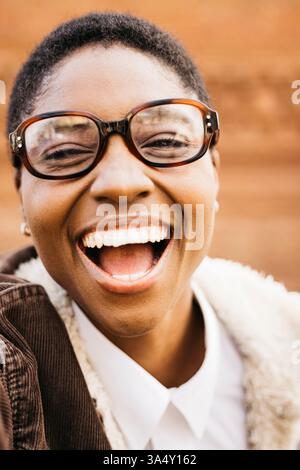 Portrait en gros plan d'une jeune femme joyeuse avec des lunettes riant du cœur sur un fond brun flou Banque D'Images