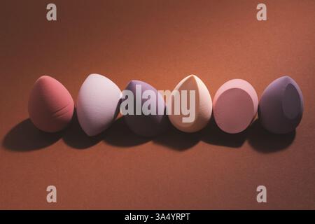 Éponges de maquillage en forme d'oeuf, mélangeurs de beauté disposés en rangée sur un fond brun. ECLAIRAGE dramatique, ombres profondes, composition élégante et artistique Banque D'Images